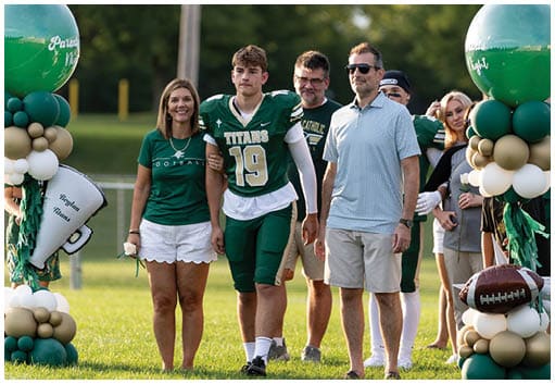 A group of people stands on a grassy field with one person wearing a green football uniform and others dressed casually near a balloon arrangement and a football. Description generated by AI