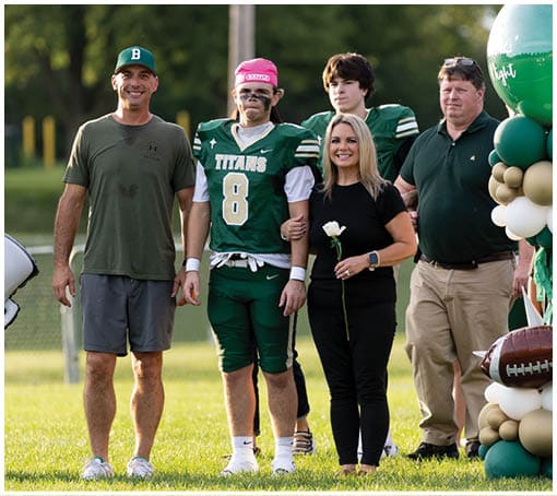 Two people stand on grass wearing athletic clothing with one person in shorts and the other in football pants, next to a large megaphone and balloons. Description generated by AI