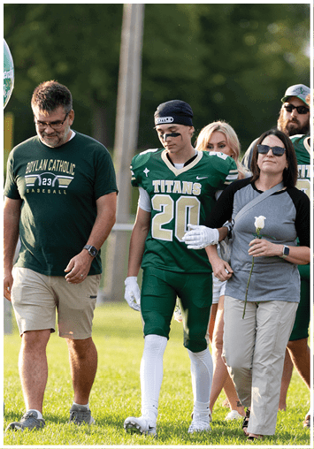 A person wearing a green football uniform with the number 20 and the word Titans walks alongside three other people, one holding a white rose, outdoors on a grassy field. Description generated by AI