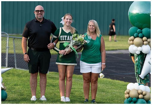 Three people stand together outdoors with one wearing a cheerleading uniform holding flowers and the others dressed casually against a green background. Description generated by AI