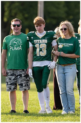 A person wearing sunglasses and a green Boylan Titans football shirt stands smiling next to a person in a green football uniform with the number 14. Description generated by AI