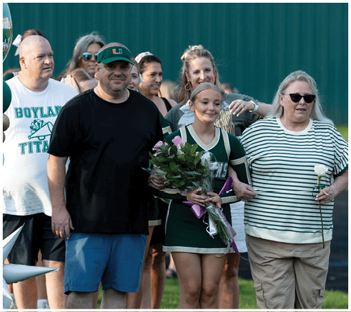 A person in a cheerleading uniform holding a bouquet of flowers is linked arm-in-arm with a person wearing sunglasses and a striped shirt, while other people stand nearby in an outdoor setting. Description generated by AI