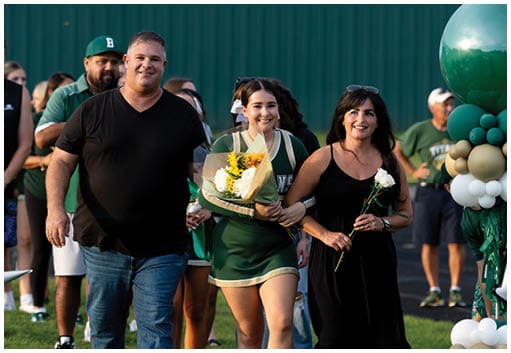 A person in a green cheerleading uniform holding a bouquet of flowers is walking arm-in-arm with another person in a black dress holding a white rose, with balloons and other people in the background. Description generated by AI