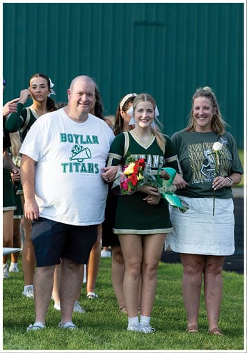 A group of people in sports attire stand on grass near a balloon decoration and a football, with one person wearing a white shirt that says Boylan Titans and another holding a bouquet of flowers. Description generated by AI