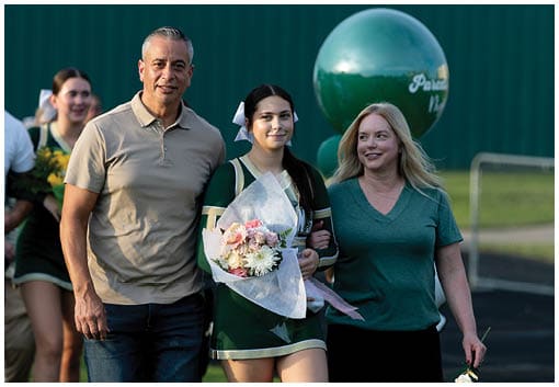 Three people stand close together in front of a dark green background with a large green balloon visible behind them. Description generated by AI
