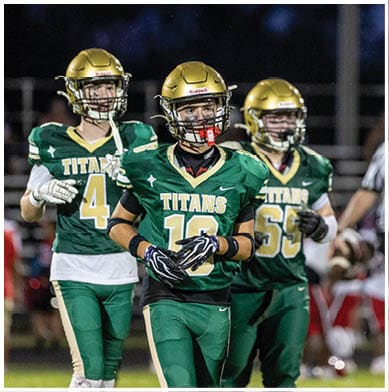 Two people in green football uniforms and gold helmets stand on a field with a referee and other players blurred in the background. Description generated by AI