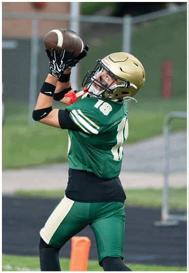 A person wearing a green football jersey with the number 18 and a gold helmet is reaching up with both hands to catch a football. Description generated by AI