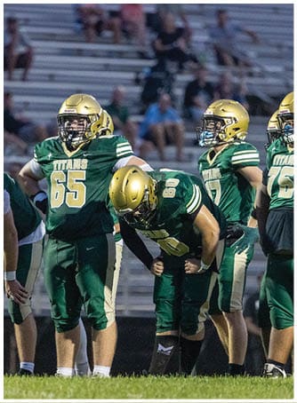 Two people in green and gold football uniforms with helmets are standing on a field with empty bleachers in the background. Description generated by AI