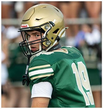 A person wearing a green football jersey with the number 19 and a gold helmet is looking over their shoulder on a sports field. Description generated by AI