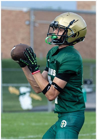 A person wearing a green sports uniform stands on a grassy field with blurred people and a fence in the background. Description generated by AI