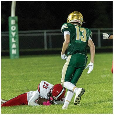 A person in a red and white football uniform is lying on the grass while another person in a green and gold football uniform with the number 13 is standing nearby on a football field. Description generated by AI