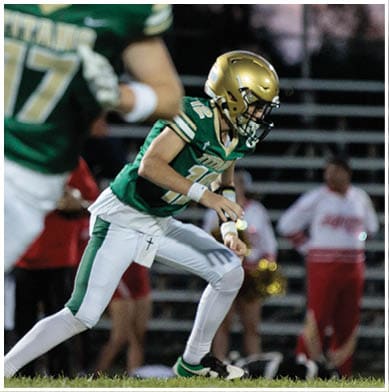 A person wearing a green and white football uniform with a gold helmet is running on a field with blurred spectators and cheerleaders in the background. Description generated by AI