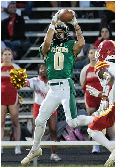 A person wearing a green and white football uniform with the number zero is jumping to catch a football while another person in a red and yellow uniform reaches toward them on a sports field with cheerleaders and spectators in the background. Description generated by AI