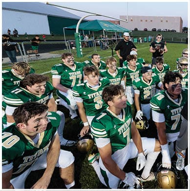 A group of athletes in green and white football uniforms with face paint are sitting and kneeling on a field near a goalpost, appearing attentive and focused. Description generated by AI