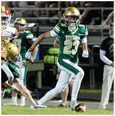 A person wearing a green and white football uniform with the number 23 is running on a field while another person in a similar uniform reaches out to grab them during a game. Description generated by AI