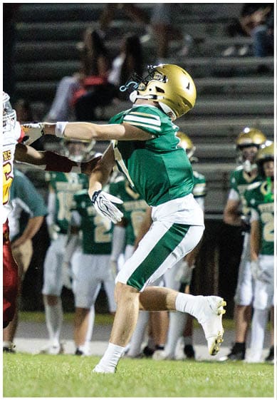 A person wearing a green and white football uniform with a gold helmet is in mid-action on a grassy field with other players and spectators blurred in the background. Description generated by AI