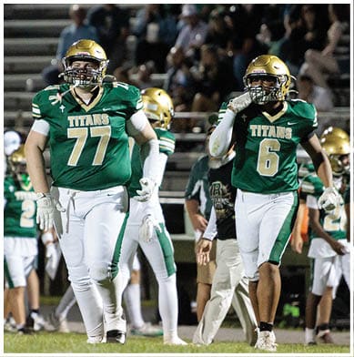 Two people in green and white football uniforms with gold helmets walk on a field during a game with spectators in the background. Description generated by AI
