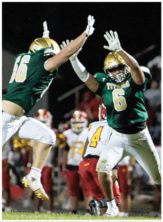 A person in a white and red football uniform is kicking a ball while two people in green and white football uniforms jump with arms raised to block the kick on a grassy field at night. Description generated by AI