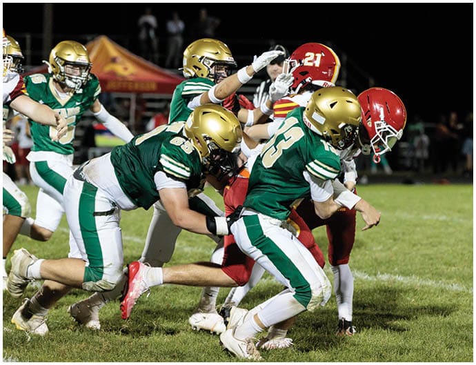 Several football players in green and gold uniforms are tackling a player in a red and yellow uniform on a grassy field during a night game. Description generated by AI