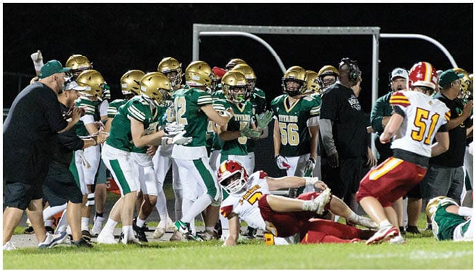 A group of football players and coaches on the sidelines watch as one player in a white and red uniform lies on the ground near another player in a white and yellow uniform during a nighttime game. Description generated by AI