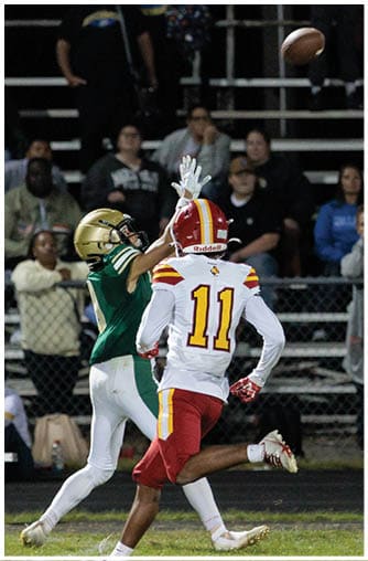 Two people in football uniforms are reaching for a football in midair on a field with spectators in the background. Description generated by AI