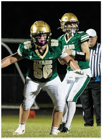 Two people wearing green and gold football uniforms are on a field at night, with one person in a protective stance and the other preparing to throw a football. Description generated by AI