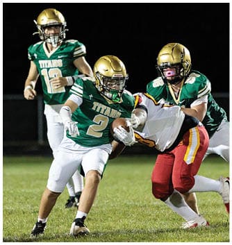Three football players in green and white uniforms with gold helmets are on a grassy field at night, with one player holding the ball while another player in red and white attempts to tackle them. Description generated by AI