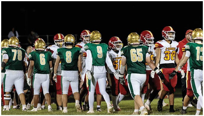 Two opposing American football teams in green and white and red and yellow uniforms are shaking hands on a field at night. Description generated by AI