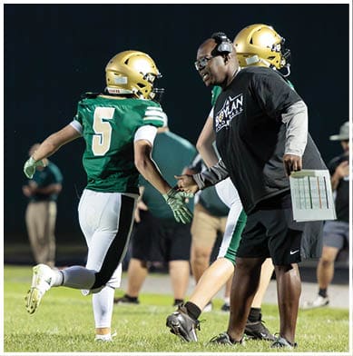 A person wearing a headset and holding a play sheet is giving a high-five to a football player in a green jersey and gold helmet on a grassy field at night. Description generated by AI
