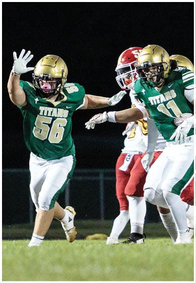 Two football players in green and gold uniforms with “TITANS" on their jerseys are actively engaged in a play against opponents in red and white uniforms on a grassy field at night. Description generated by AI