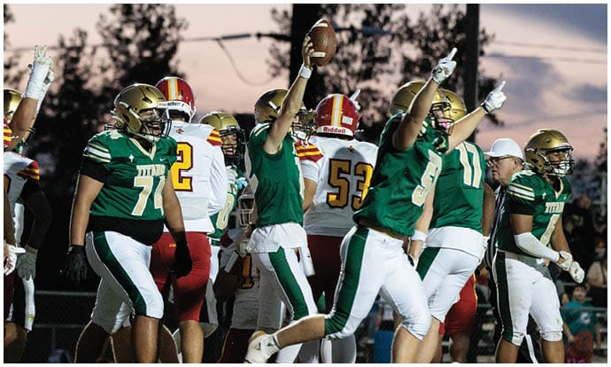 People wearing green and white football uniforms celebrate on a field at dusk while players in red and white uniforms stand nearby. Description generated by AI