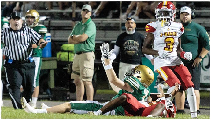 Two football players are on the ground during a game while a referee signals and other people watch from the sidelines. Description generated by AI