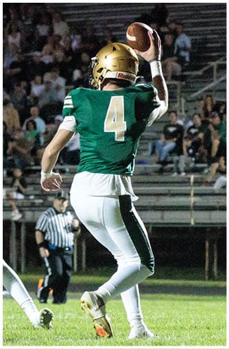 A person wearing a green football jersey with the number 4 and a gold helmet is preparing to throw a football on a grassy field with a referee and spectators in the background. Description generated by AI