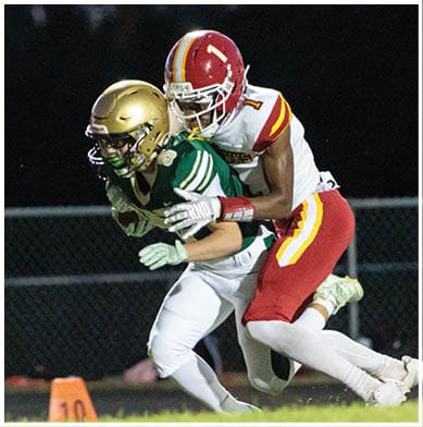 Two people in football uniforms are engaged in a tackle on a field at night, with one wearing a green and gold uniform and the other in a white and red uniform. Description generated by AI