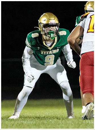 A person in a green and white football uniform with the number 9 is crouched in a ready stance on a grassy field during a nighttime game. Description generated by AI