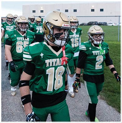 A group of people wearing green football uniforms and gold helmets walk together on a gravel path near a grassy area and a chain-link fence. Description generated by AI