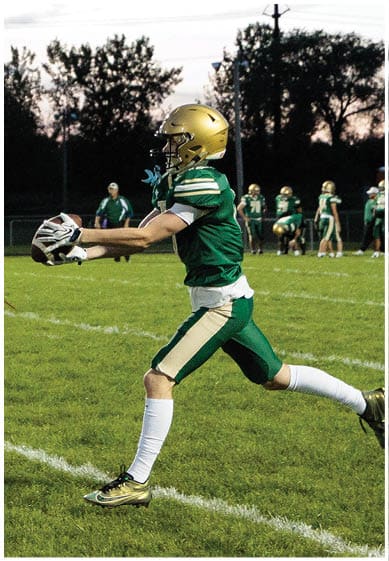 A person in a green and gold football uniform is catching a football while running on a grassy field during a game at dusk. Description generated by AI