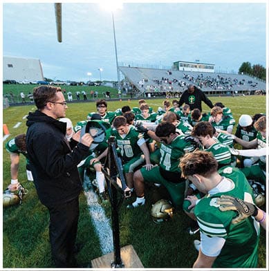 A group of people in green sports uniforms are gathered on a football field with two adults standing nearby, one holding a cap, during a moment of pause or discussion. Description generated by AI