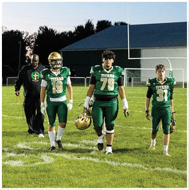 Two people wearing green football uniforms with helmets in hand walk on a grassy field near a goalpost. Description generated by AI