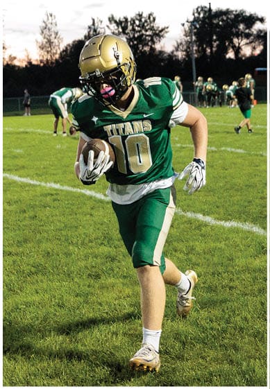 A person in a green and white football uniform is running on a grassy field with other people and a bus visible in the background during dusk. Description generated by AI