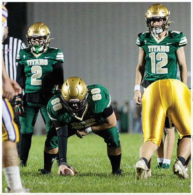 Two people in green football uniforms with gold helmets are positioned on a field, with one preparing to snap the ball and the other standing behind. Description generated by AI