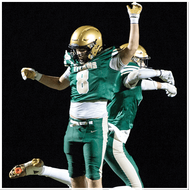 A person wearing a green football jersey with the number 8 and a gold helmet is raising their arms in celebration during a nighttime game. Description generated by AI