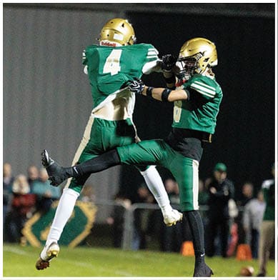 Two people in green and gold football uniforms are jumping and reaching for a football during a nighttime game with blurred spectators in the background. Description generated by AI