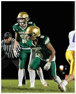 Two football players in green and gold uniforms and a referee in a black and white striped shirt are positioned on a field under bright lights. Description generated by AI