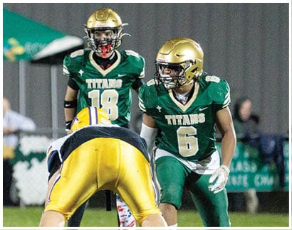 Three football players in green and gold uniforms are positioned on a grassy field during a game, with one player in yellow and blue bent over in front of them. Description generated by AI