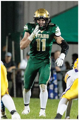 A person wearing a green football uniform with the number 11 and a gold helmet is standing on a grassy field, appearing to signal or communicate during a game. Description generated by AI