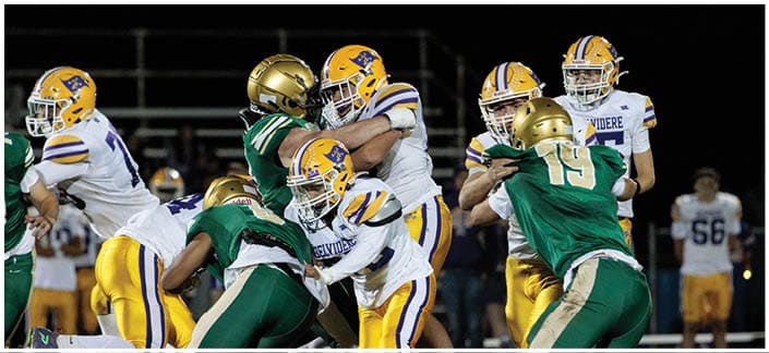 Players in yellow and white uniforms and players in green uniforms are engaged in a football game on a field at night. Description generated by AI