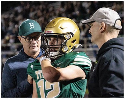 A person wearing a gold football helmet and green jersey is being assisted by another person wearing glasses and a green cap with a white letter B. Description generated by AI