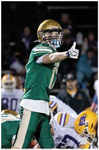 A person wearing a green and gold football uniform and helmet is pointing forward on a football field during a game. Description generated by AI