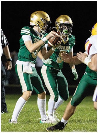 A person in a green football uniform is holding a football and preparing to pass while another person in a white and yellow football uniform is approaching. Description generated by AI